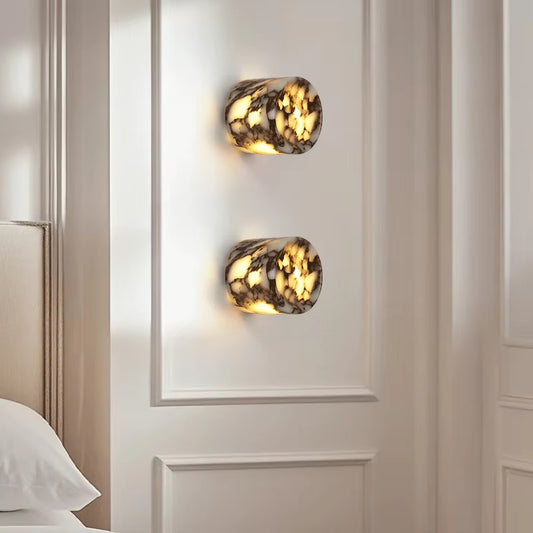Two marble cylinder wall sconces, lit, one above the other on a panelled wall beside a bed.