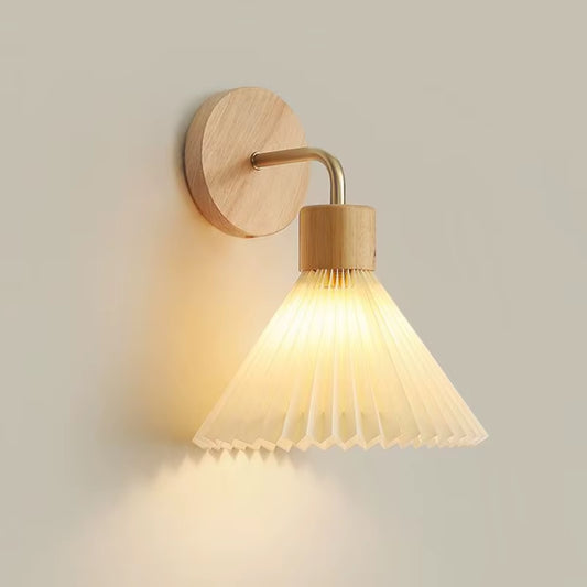 Wall sconce with a white pleated cone shade on a light wood plate, lit, on a plain wall.