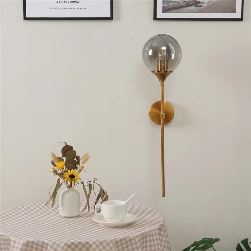 Smoke gray glass globe wall sconce on a gold-color metal stem beside a small table with flowers.