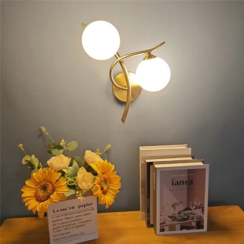 Two-globe wall sconce with frosted glass globes, lit, above sunflowers and books on a wood table.