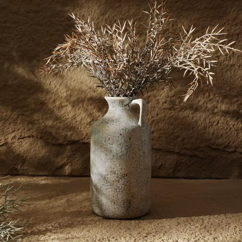Tall ceramic jug vase holding dried branches against a textured wall.