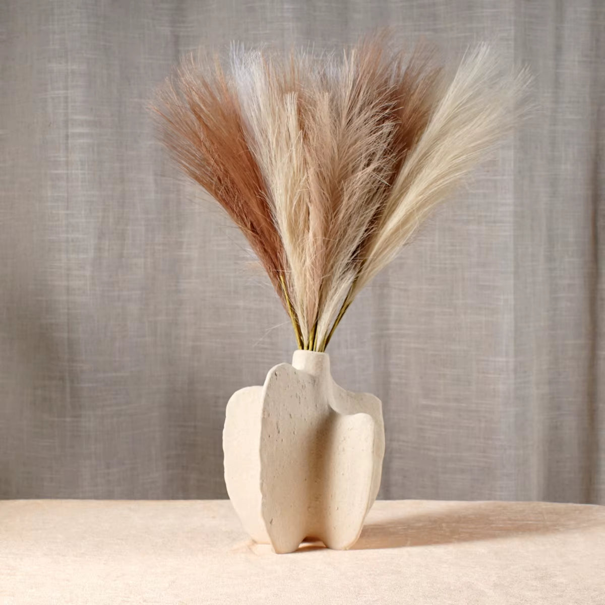 White cleft block ceramic vase holding dried pampas grass in front of a linen curtain.