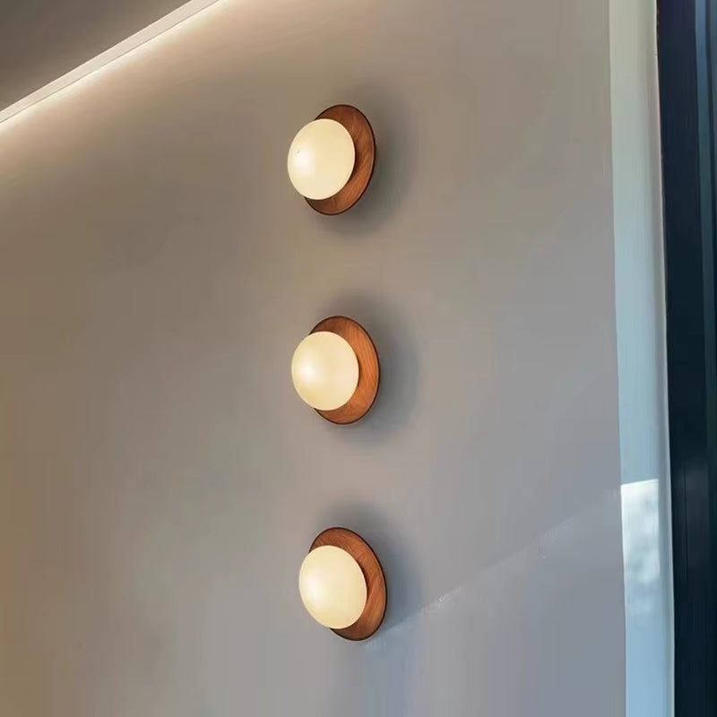 Three walnut-tone wood disc wall sconces with glass globes, lit, in a row on a wall.