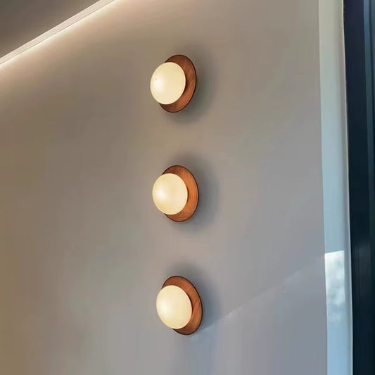 Three walnut-tone wood disc wall sconces with glass globes, lit, in a row on a wall.