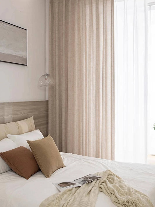 Natural beige semi-sheer curtain panel hanging beside a bed with neutral bedding.