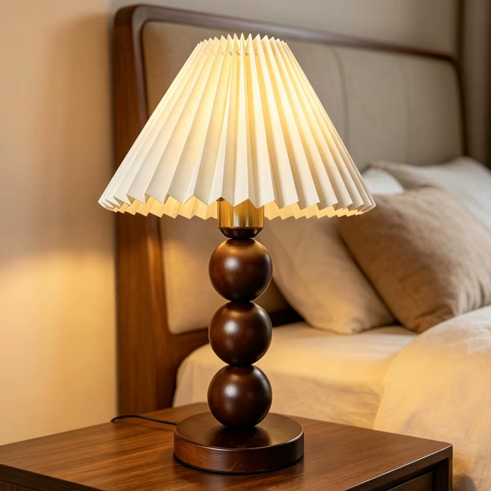 Walnut-color stacked-sphere table lamp with a white pleated shade, lit, on a bedside table.